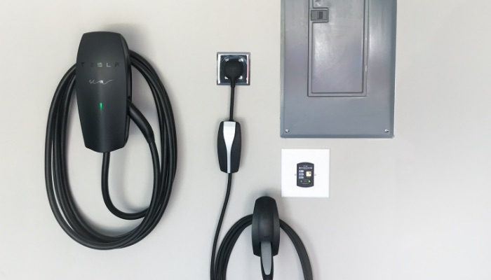 A Tesla Wall Connector with a coiled cable mounted in a garage beside the electrical panel and a second charging connector.